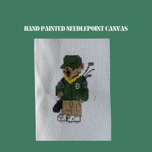 May include: Hand-painted needlepoint canvas featuring a golfer in a green hat, sweater, and khaki pants, carrying golf clubs. The letter "B" is on the sweater. The background is white with a green border. Text at the top reads "HAND PAINTED NEEDLEPOINT CANVAS."