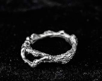 May include: A silver ring with a textured, organic design. The ring has a rough, bark-like appearance, with an open, irregular shape. The ring is set against a black background, highlighting its intricate details and metallic sheen.
