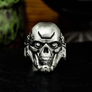 May include: A silver-colored skull ring with a detailed, mechanical design. The ring features a skull with visible eye sockets, teeth, and intricate metalwork, creating a gothic or cyberpunk aesthetic. The ring is likely made of metal.
