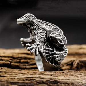 Puede incluir: Un anillo plateado detallado con un diseño de rana. La rana está esculpida con piel texturizada y detalles intrincados, incluyendo ojos visibles y patas palmeadas. El anillo está sobre un fondo oscuro y una superficie de madera.