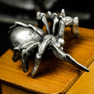 May include: A detailed, silver-colored spider figurine with eight legs and a textured body. The arachnid is posed on a wooden surface, showcasing its intricate design and metallic sheen. The figurine is a decorative item.