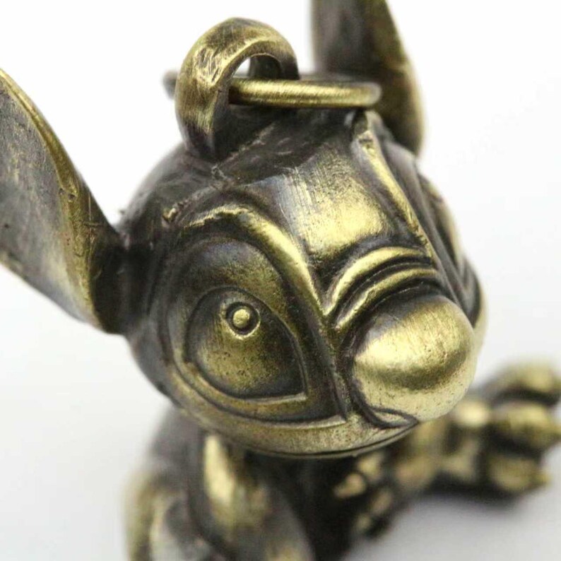 May include: A bronze-colored figurine of the character Stitch from the animated film Lilo & Stitch. The figurine has large ears, a prominent nose, and a loop for attachment. The metal has a slightly aged appearance.
