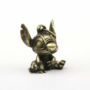 May include: A bronze-colored Stitch figurine in a seated position. The figure has large ears, a wide nose, and a loop for attaching to a keychain. The figurine is detailed with visible facial features and body contours.