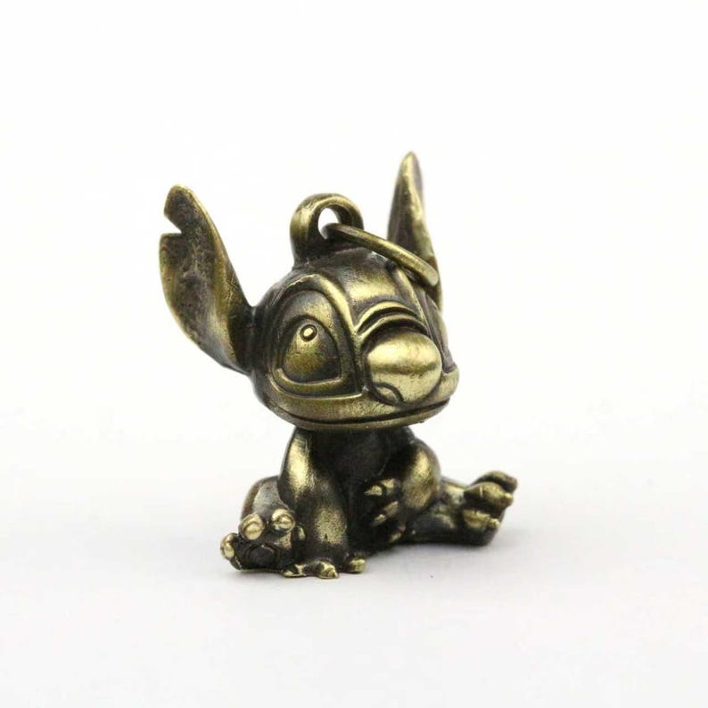 May include: A bronze-colored Stitch figurine in a seated position. The figure has large ears, a wide nose, and a loop for attaching to a keychain. The figurine is detailed with visible facial features and body contours.