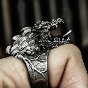 May include: A silver ring with a detailed dragon head design. The dragon's mouth is open, showing sharp teeth and a tongue. The ring has a textured band and a detailed, three-dimensional dragon head.