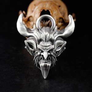 May include: A detailed silver pendant featuring a demonic face with horns, a pointed chin, and a ring for a chain. The pendant is set against a blurred background of a skull. The pendant is a gothic style accessory.