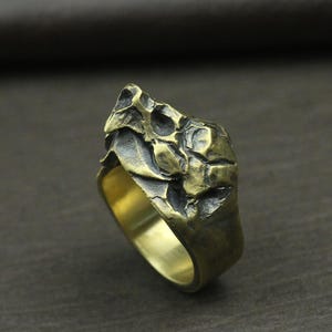 May include: A bronze-colored ring with a textured, rugged design. The ring's surface has an uneven, rocky appearance, with a dark, aged patina. The band is smooth and polished, contrasting with the textured top. A unique piece of jewelry.