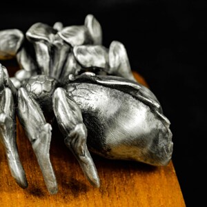May include: A detailed, silver-colored spider sculpture with intricate detailing. The spider is positioned on a wooden surface, with a dark background. The sculpture's legs and body are clearly defined, showcasing the craftsmanship.