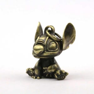 May include: A bronze-colored Stitch figurine, sitting with its arms and legs outstretched. The figurine has large ears, a round head, and a key ring attached to the top. The figure is detailed with visible toes and a smiling expression.