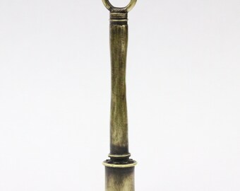 May include: A vintage-style bottle stopper with a brass finish. The stopper features a cylindrical base, a long, slender stem, and a circular handle at the top. The metal has an aged appearance, suggesting it is an antique or a replica.