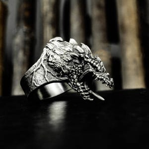 May include: A silver ring with a detailed dragon head design. The dragon's head is sculpted with textured scales, an open mouth, and sharp teeth. The ring band is smooth and polished.