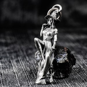 Könnte beinhalten: Silberfarbener Anhänger, der eine sitzende Hexenfigur mit spitzem Hut und langem Kleid zeigt. Die Hexe sitzt auf einem dunklen, strukturierten Felsen. Der Anhänger hat oben eine Schlaufe für eine Kette. Der Hintergrund ist ein verschwommenes, dunkles Grau.