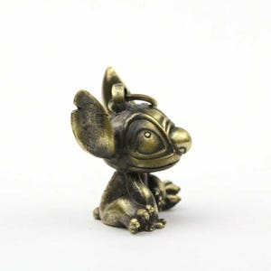 May include: A bronze-colored figurine of the character Stitch from the Disney movie Lilo & Stitch. The figurine is in a seated position with large ears and a detailed facial expression. The keychain is made of metal.