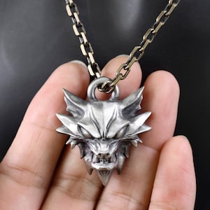 May include: A silver-toned metal pendant shaped like a stylized wolf head with pointed ears and bared teeth. The pendant is attached to a thick, dark-colored metal chain with rectangular links, featuring a weathered, matte finish throughout.