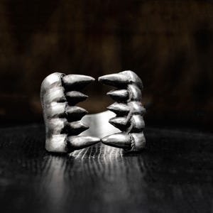 May include: A silver-toned ring with a unique design. The ring features two opposing claw-like structures, each with multiple pointed tips. The ring's design is detailed and has a textured appearance, set against a dark background.