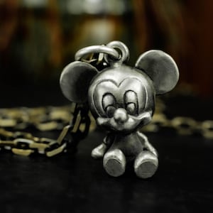 以下が含まれることがあります： 座っているミッキーマウスの姿が描かれた、シルバーカラーのペンダントネックレス。ペンダントは、大きな耳、笑顔の顔、チェーンを備えた詳細な3次元デザインです。このネックレスはディズニーファンに適しています。