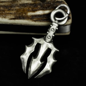 May include: A silver-colored pendant with a trident design. The pendant has a ring at the top for attaching to a chain. The trident has sharp, pointed tines and a textured surface. The pendant is likely made of metal and is displayed against a dark background.
