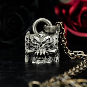 May include: A silver-colored pendant necklace featuring a detailed skull design. The pendant is square-shaped with a skull face on the front, a hinged top, and a chain. The background is dark with a blurred red rose.