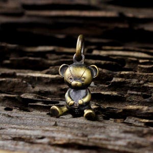 Puede incluir: Un pequeño dije de oso de peluche de latón de estilo antiguo con un lazo para colgar. El oso tiene la cabeza cosida y está sentado con las piernas estiradas. El dije es de color latón oscuro.