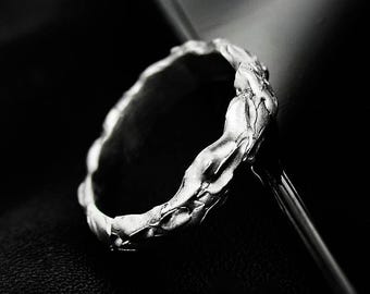 May include: A silver ring with a textured, organic design. The ring has an uneven surface, with a rough, almost bark-like texture. The ring is set against a dark background, highlighting its metallic sheen.