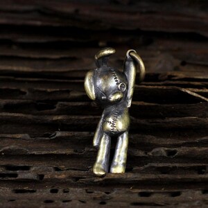 May include: A small, bronze-colored metal charm in the shape of a teddy bear. The bear has stitched details, a single eye, and is raising one arm. The charm is designed with a loop for attachment.