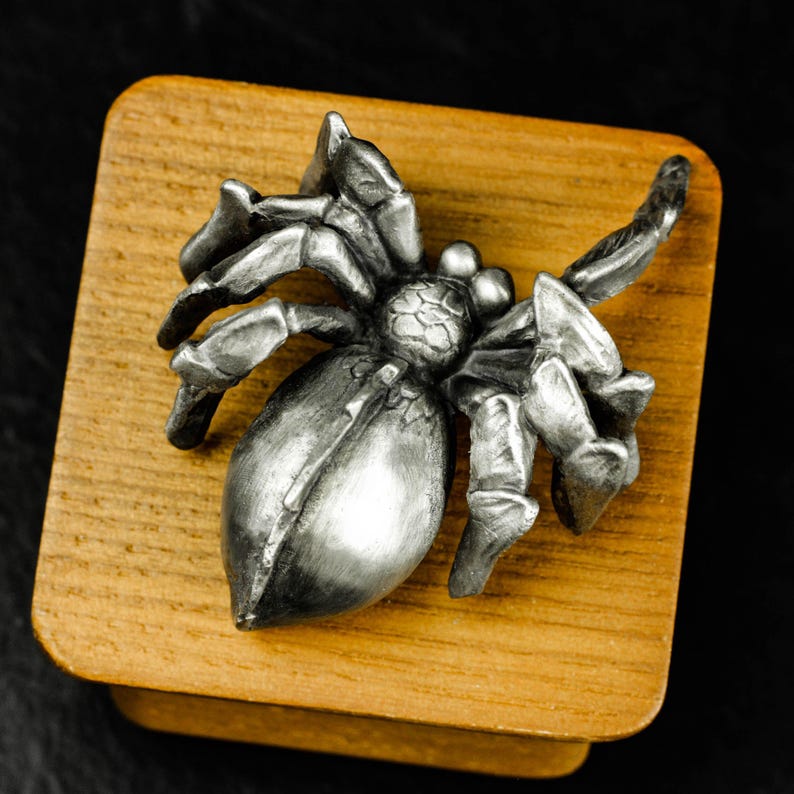 May include: A detailed, silver-colored spider figurine with eight legs, mounted on a small, square wooden block. The spider's body is bulbous, and its legs are articulated. The wooden block has a natural wood grain finish, and the background is black.