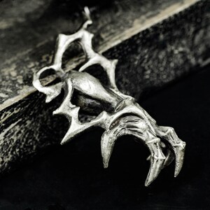 May include: A silver-toned pendant in the shape of a claw. The claw has sharp, pointed tips and a detailed, textured surface. The pendant is likely made of metal and has a gothic or fantasy theme.