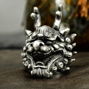 Può includere: Un anello argentato dettagliato raffigurante la testa di una creatura mitica. L'anello presenta dettagli intricati, tra cui una bocca aperta, denti affilati e tratti del viso elaborati. Il design include corna ed elementi decorativi.