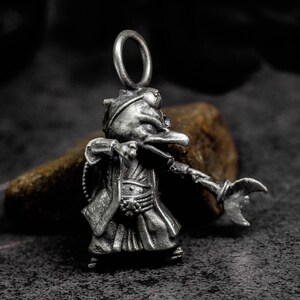 May include: A silver-colored pendant in the shape of a stylized figure with a long beak, wearing a robe and hat, holding a crescent-shaped weapon. The pendant has a loop at the top for attaching to a chain or cord.