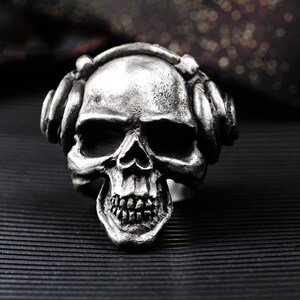 Könnte beinhalten: Ein silberner Totenkopfring mit Kopfhörern. Der Schädel hat detaillierte Zähne und Augenhöhlen. Die Kopfhörer sind an den Schädel angepasst, was ein einzigartiges Design ergibt. Der Ring ist vor einem dunklen Hintergrund platziert.