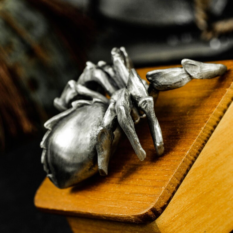 May include: A detailed, silver-colored spider figurine with intricate leg and body details. The spider is positioned on a wooden surface, showcasing its textured appearance. The figurine's design suggests a focus on arachnid-themed decor or collectibles.