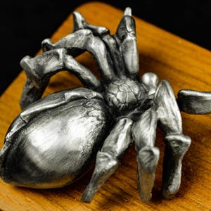 May include: A detailed, silver-colored spider sculpture with eight legs and a textured body. The arachnid is positioned on a wooden surface, showcasing its intricate design and metallic sheen. The sculpture's legs are bent, giving it a lifelike appearance.