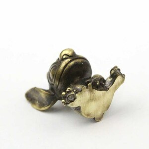 May include: A small, bronze-colored frog figurine with detailed features. The frog is depicted in a relaxed pose, with its front legs extended. The figurine has a smooth, polished finish and is set against a white background.