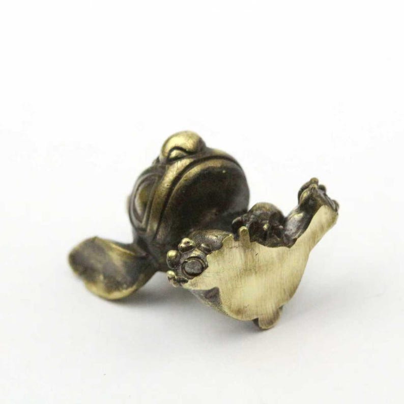 May include: A small, bronze-colored frog figurine with detailed features. The frog is depicted in a relaxed pose, with its front legs extended. The figurine has a smooth, polished finish and is set against a white background.