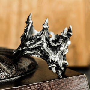 May include: A silver-colored ring with a crown design. The ring features a detailed, textured surface with multiple pointed spikes. The ring is set against a dark wooden surface with a blurred background.