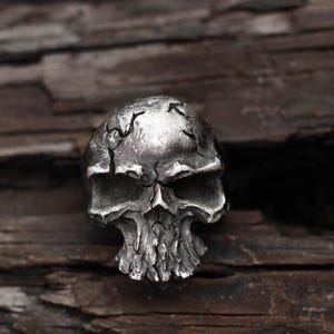 May include: A detailed, silver-colored skull ring with a cracked, weathered appearance. The ring features a realistic skull design, showcasing the eye sockets, nasal cavity, and teeth. The ring is likely made of metal and is a gothic or biker-style accessory.