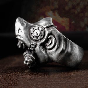 Puede incluir: Un anillo plateado con un diseño detallado de máscara. El anillo presenta intrincados elementos mecánicos y de calavera, incluyendo un detalle circular en forma de engranaje y una pequeña calavera. El anillo tiene una apariencia texturizada y envejecida.
