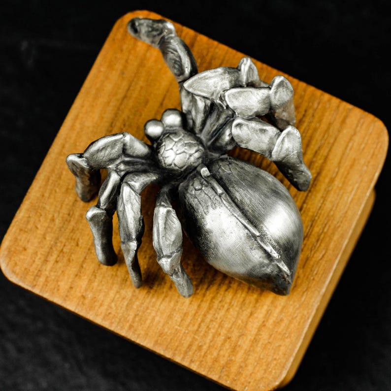 May include: A detailed, silver-colored spider figurine with eight legs, positioned on a square, wooden block. The spider's body has intricate detailing, and the wood has a natural grain. The background is dark, emphasizing the spider and the wood.