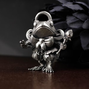 May include: A detailed silver-toned frog figurine, adorned with a bow tie, ruffled collar, and a staff. The frog has a loop at the top, suggesting it could be a pendant or charm. The figure is standing on a dark surface.