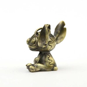 May include: A bronze-colored figurine of the Disney character Stitch, in a seated pose. The figurine has large ears, a round head, and a loop for attachment. The details include etched patterns and a weathered finish, suggesting an antique or vintage style.