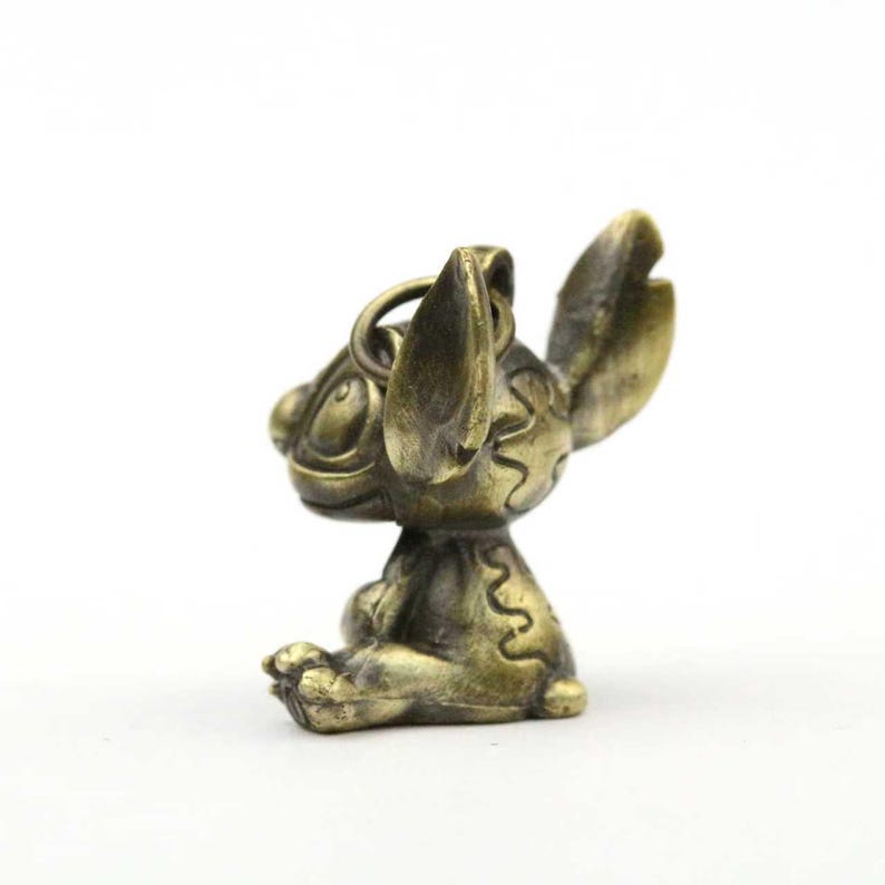 May include: A bronze-colored figurine of the Disney character Stitch, in a seated pose. The figurine has large ears, a round head, and a loop for attachment. The details include etched patterns and a weathered finish, suggesting an antique or vintage style.