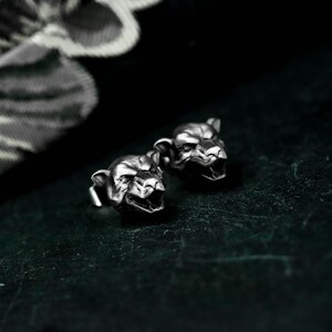 Könnte beinhalten: Ein Paar silberfarbene Ohrstecker. Jeder Ohrring ist in Form eines knurrenden Pantherkopfes mit detaillierten Gesichtszügen geformt. Die Ohrringe sind vor einem dunklen, strukturierten Hintergrund platziert, der den metallischen Glanz hervorhebt.