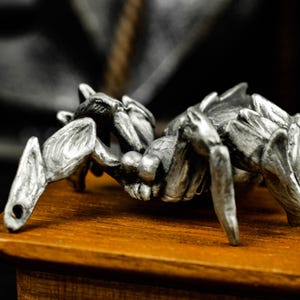 May include: A detailed, silver-colored spider figurine with multiple legs and a textured body. The arachnid is positioned on a wooden surface, showcasing its intricate design. The background is blurred, emphasizing the spider's metallic appearance.