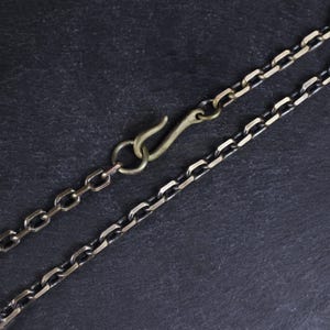 May include: Close-up of a necklace with a hook clasp. The chain is made of interlocking links in a dark silver color, with a brass-colored hook. The background is a dark, textured surface.