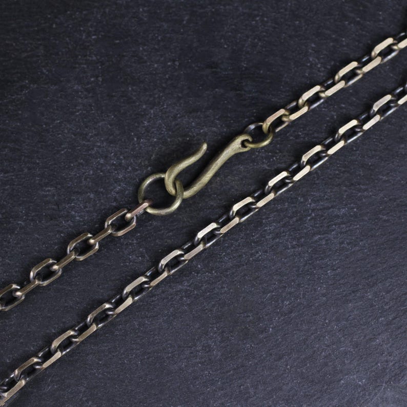 May include: Close-up of a necklace with a hook clasp. The chain is made of interlocking links in a dark silver color, with a brass-colored hook. The background is a dark, textured surface.