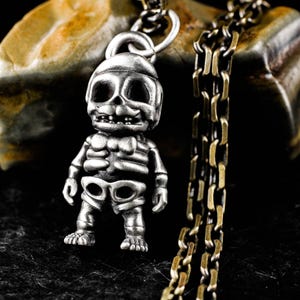 May include: A silver-colored pendant necklace featuring a stylized skeleton figure. The pendant is detailed with a skull-shaped head, rib cage, and shorts. The necklace has a silver chain and a contrasting gold-toned chain.