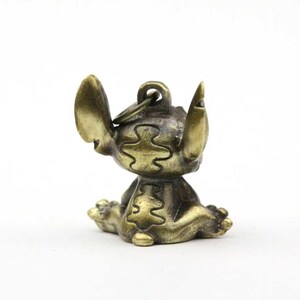 May include: A bronze-colored figurine of the character Stitch from the animated film Lilo & Stitch. The figurine is in a seated position with its arms raised. The figure has a key ring attached to the top of its head.