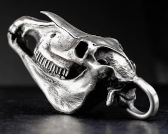 May include: A detailed, silver-toned pendant in the shape of a horse skull. The skull features intricate detailing of the teeth, eye sockets, and nasal cavity. A loop is attached for hanging. The pendant is set against a dark background.