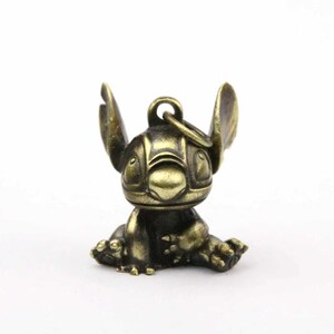 May include: A bronze-colored Stitch figurine, featuring large ears and a seated pose. The keychain-style charm has a ring at the top for attachment. The figure is detailed with etched facial features and a textured finish.
