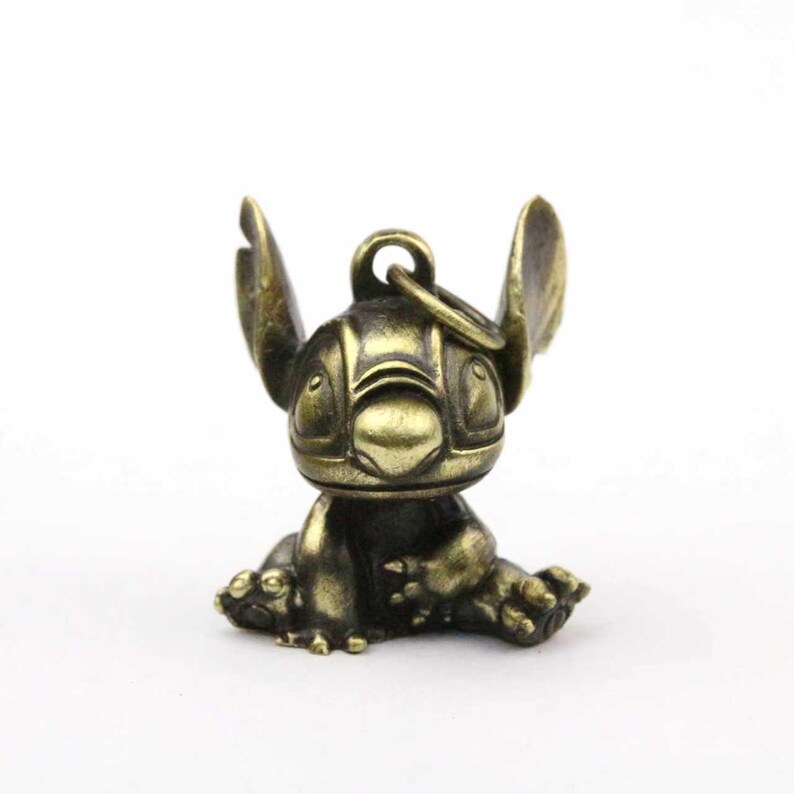 May include: A bronze-colored Stitch figurine, featuring large ears and a seated pose. The keychain-style charm has a ring at the top for attachment. The figure is detailed with etched facial features and a textured finish.